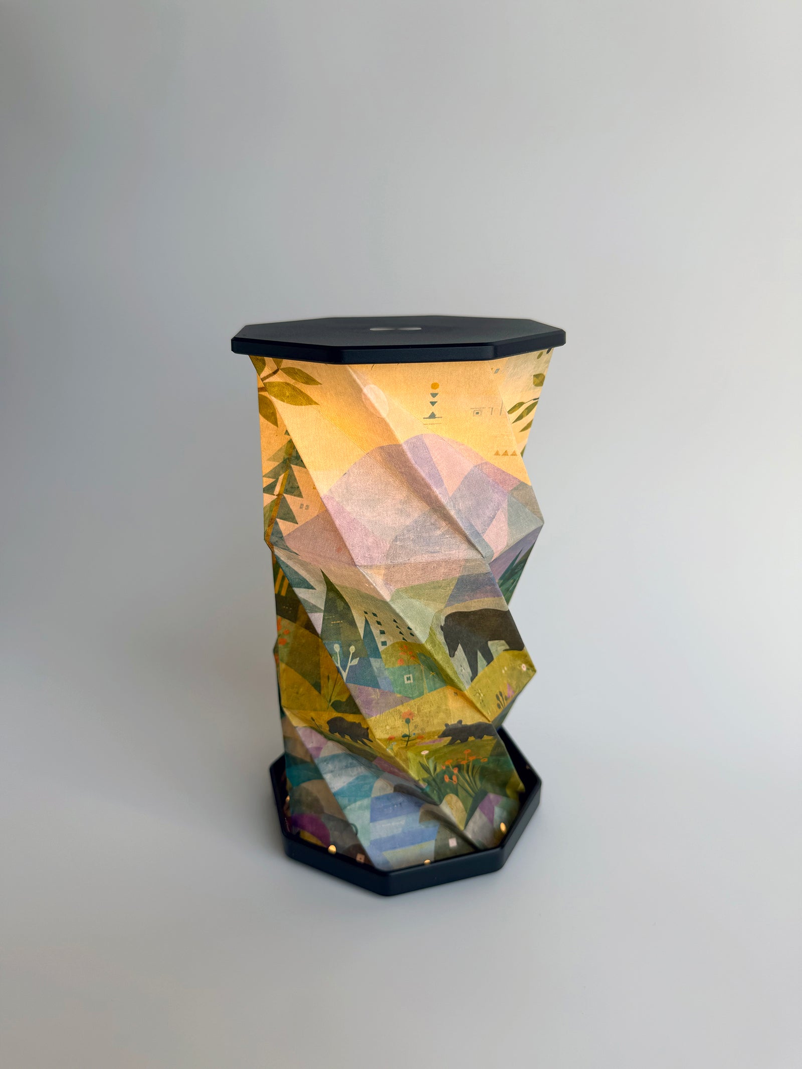 An octagonal-shaped lamp featuring a faceted, twist and pull design covered in an abstract, geometric art print. The print depicts a family of black bears roaming through a hillside meadow scene with a blue stream and mountain in the background, in tones of blue, purple, green and yellow, set against a dark charcoal-black base and top surface, photographed on a clean white studio background.