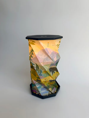 An octagonal-shaped lamp featuring a faceted, twist and pull design covered in an abstract, geometric art print. The print depicts a family of black bears roaming through a hillside meadow scene with a blue stream and mountain in the background, in tones of blue, purple, green and yellow, set against a dark charcoal-black base and top surface, photographed on a clean white studio background.