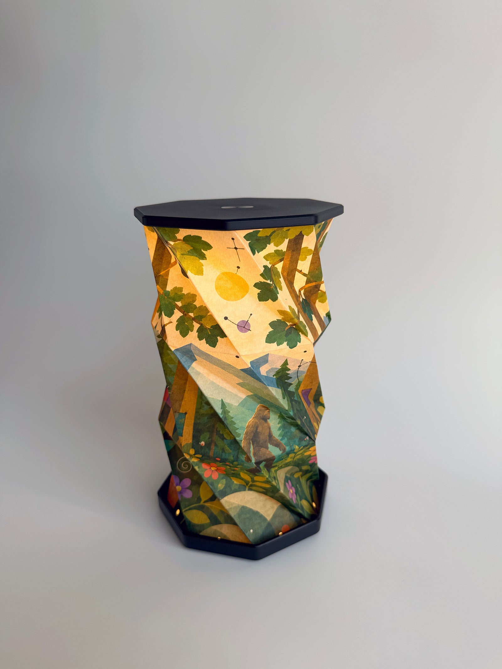 An octagonal-shaped lamp featuring a faceted, twist and pull design covered in an abstract, geometric art print. The print depicts Bigfoot, or Sasquatch, walking in a forest scene, with small flowers in the foreground and evergreen trees and mountains in the background, set against a dark charcoal-black base and top surface, photographed on a clean white studio background.