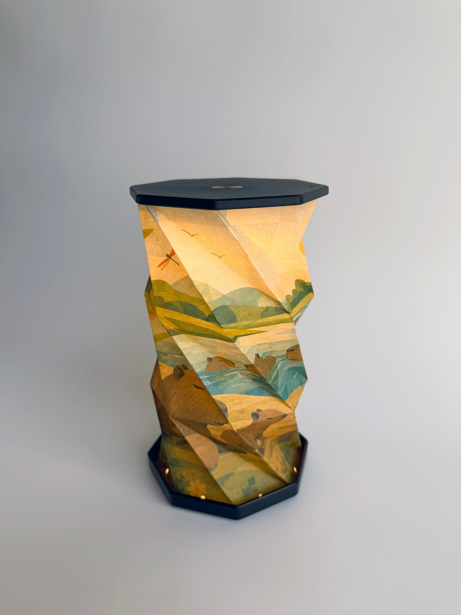 An octagonal-shaped lamp featuring a faceted, twist and pull design covered in an abstract, geometric art print. The print depicts Capybara animals sunbathing and swimming in a small lake with rolling hills and mountain motifs in greenish blue earth tones with dragonflies and flying bird, set against a dark charcoal-black base and top surface, photographed on a clean white studio background.