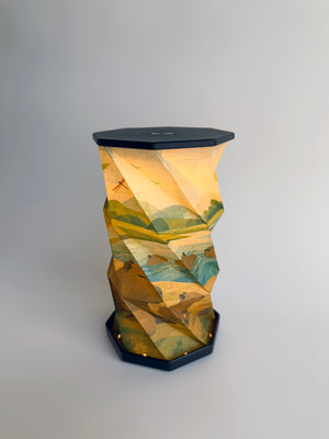 An octagonal-shaped lamp featuring a faceted, twist and pull design covered in an abstract, geometric art print. The print depicts Capybara animals sunbathing and swimming in a small lake with rolling hills and mountain motifs in greenish blue earth tones with dragonflies and flying bird, set against a dark charcoal-black base and top surface, photographed on a clean white studio background.
