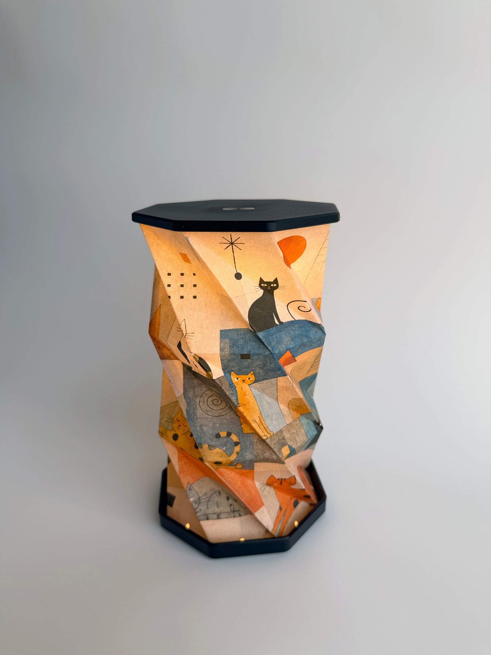 An octagonal-shaped lamp featuring a faceted, twist and pull design covered in an abstract, geometric art print. The print depicts many different cats sitting and lounging peacefully while looking at the viewer, with small abstract houses  and archway motifs in warm tones of brown, beige and orange, set against a dark charcoal-black base and top surface, photographed on a clean white studio background.