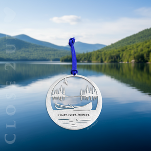 A stainless steel keepsake ornament with a silhouette of an eagle soaring over a lake scene and Canoe with trees in the background, with the text 'Enjoy, Every Moment’, strung with a faux suede cord. Shown in front of a photo of a mountain lake scene.