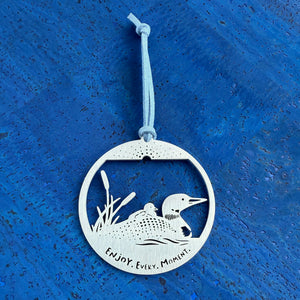 A stainless steel keepsake ornament with a swimming Loon and chick on its back,  with cattail in the background, and the text 'Enjoy, Every Moment’, strung with a faux suede cord.