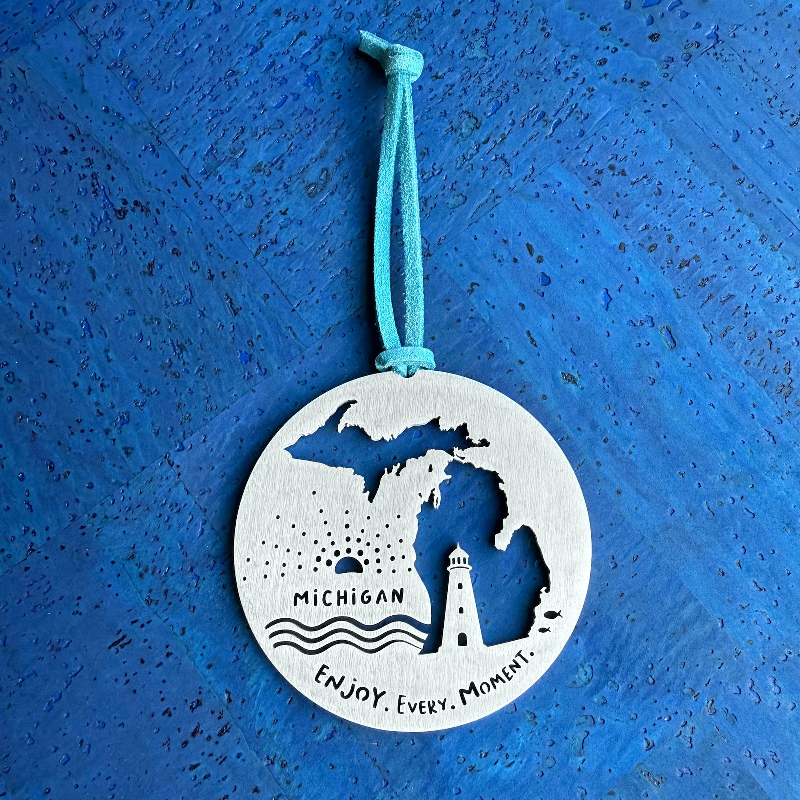 A round stainless steel keepsake ornament with a cut out map of the state of Michigan, with the text 'Michigan' and 'Enjoy, Every Moment’, accented with a lighthoust, small waves and a sunset, strung with a faux suede cord.