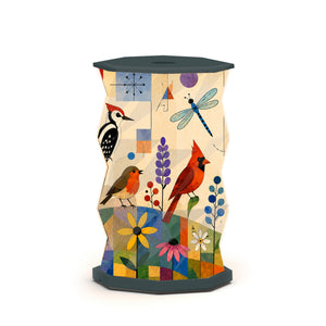 A sculptural, hexagonal side table or lamp featuring a faceted, twisted design covered in an abstract, geometric art print. The print depicts birds and flower motifs in warm earth tones of beige, set against a dark charcoal/black base and top surface, photographed on a clean white studio background.