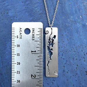 A stainless steel rectangular-shaped necklace pendant featuring a cut-out shape of Lake Champlain, with wave curl accents, by Close 2 UR Heart, shown alongside a metal ruler for size comparison.