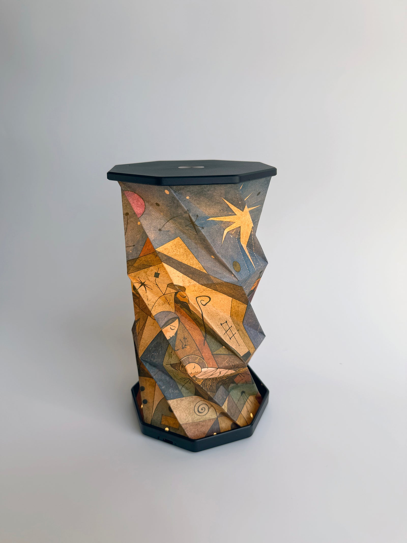 An octagonal-shaped lamp featuring a faceted, twist and pull design covered in an abstract, geometric art print. The print depicts Biblical figures of the Nativity scene and star-like motifs in warm earth tones of brown, beige, and orange, set against a dark charcoal-black base and top surface, photographed on a clean white studio background.