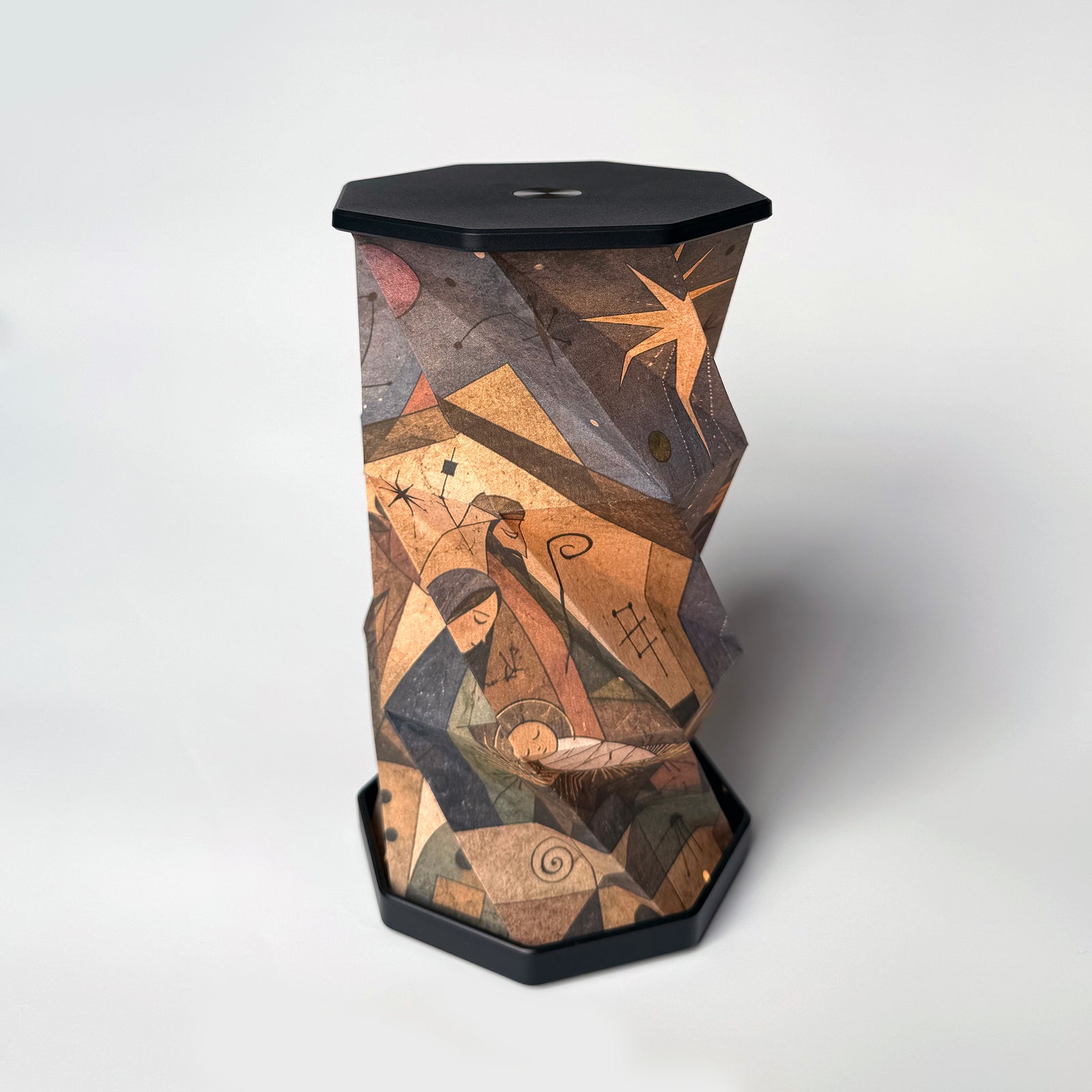 A sculptural, hexagonal side table or lamp featuring a faceted, twisted design covered in an abstract, geometric art print. The print depicts figures and star-like motifs in warm earth tones of brown, beige, and orange, set against a dark charcoal/black base and top surface, photographed on a clean white studio background.