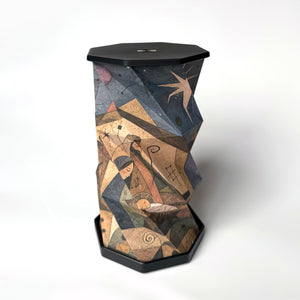 A sculptural, hexagonal side table or lamp featuring a faceted, twisted design covered in an abstract, geometric art print. The print depicts figures and star-like motifs in warm earth tones of brown, beige, and orange, set against a dark charcoal/black base and top surface, photographed on a clean white studio background.