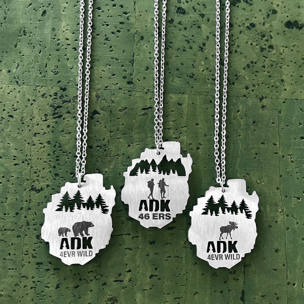 Stainless steel necklace pendants in the shape of the Adirondack Park, NY, featuring bears, moose and hiker with trees in the background, with the text "ADK 4EVR WILD".