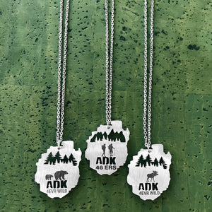 Stainless steel necklace pendants in the shape of the Adirondack Park, NY, featuring bears, moose and hiker with trees in the background, with the text "ADK 4EVR WILD".