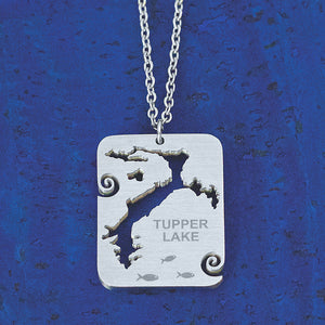 A stainless steel necklace pendant featuring a cut out of Tupper Lake, New York with small wave curl accents, and the text "Tupper Lake", by Close 2 UR Heart.