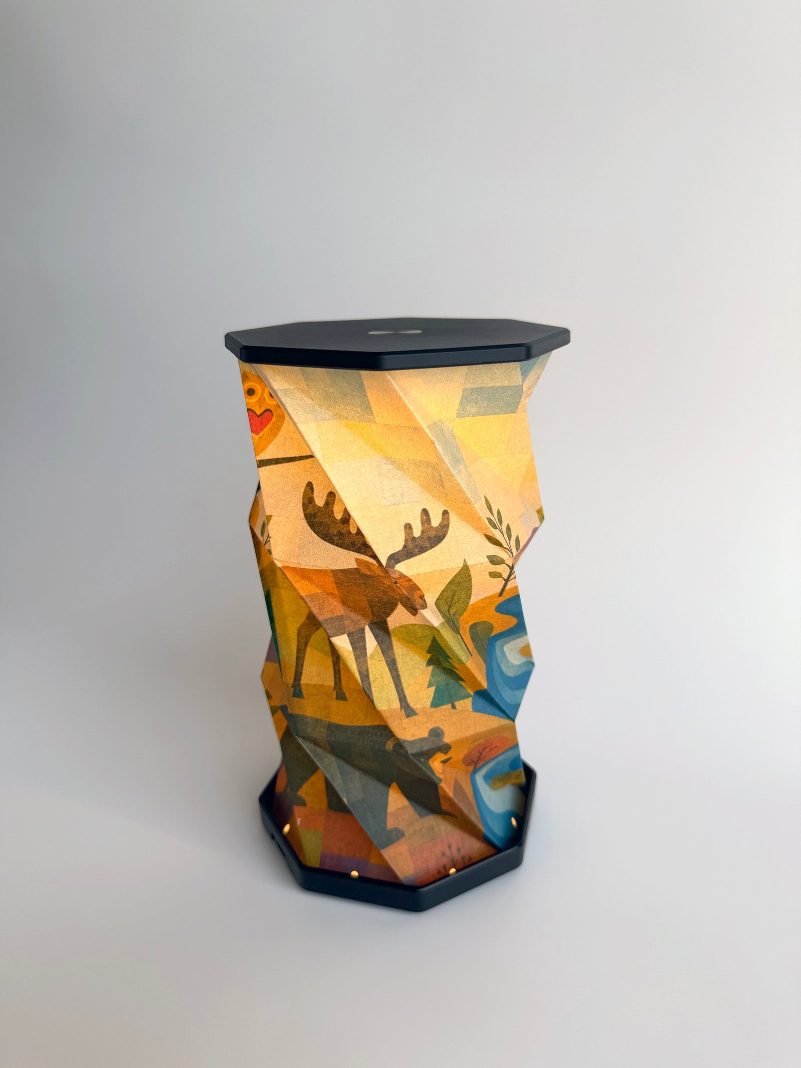 An octagonal-shaped lamp featuring a faceted, twist and pull design covered in an abstract, geometric art print. The print depicts an outdoor woodland scene with a variety of animals such as moose, bear, owl and fish in a stream, with small trees on rolling hills in warm earth tones of brown, beige, and orange, set against a dark charcoal-black base and top surface, photographed on a clean white studio background.